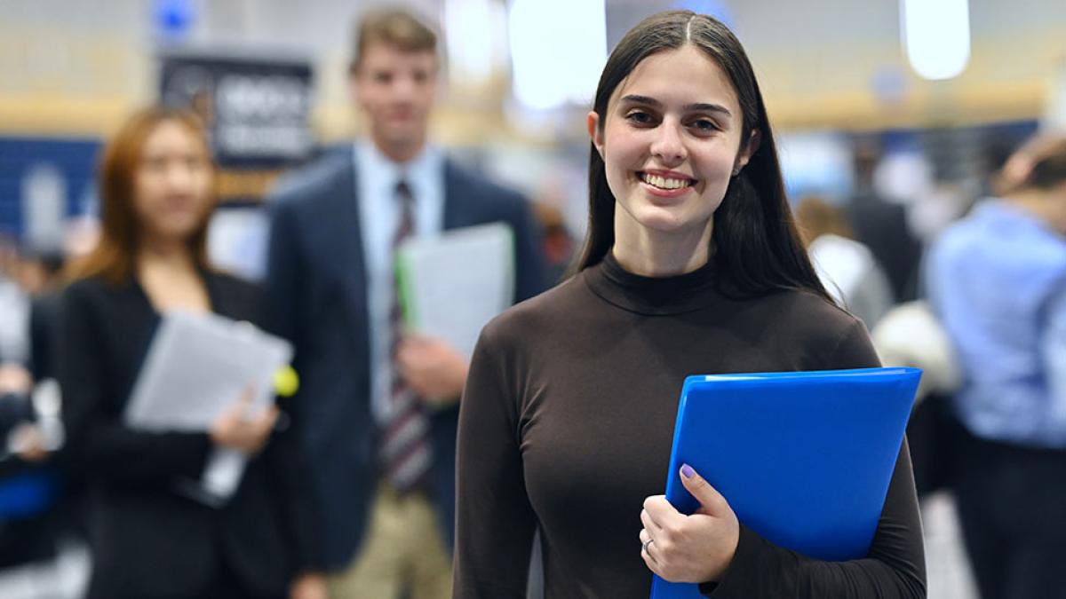 A Pace student holds a blue folder and smiles, a career fair behind her