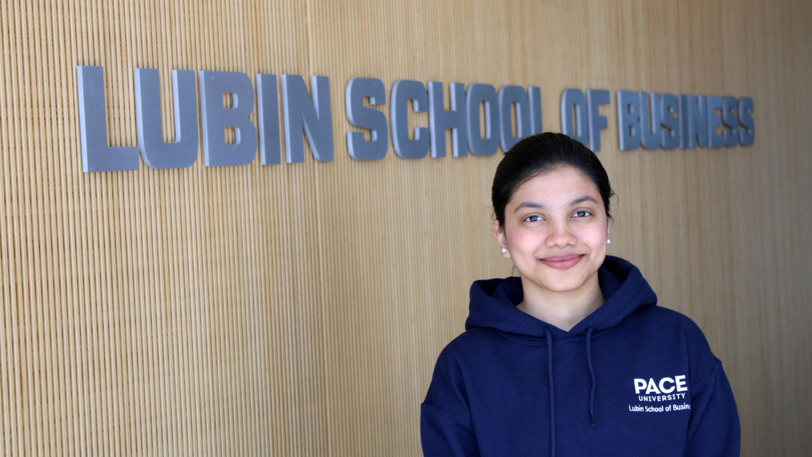 Lubin student Shruti Dhapodkar '23 standing in front of the Lubin School of Business sign on the 4th Floor of One Pace Plaza on the New York City Campus