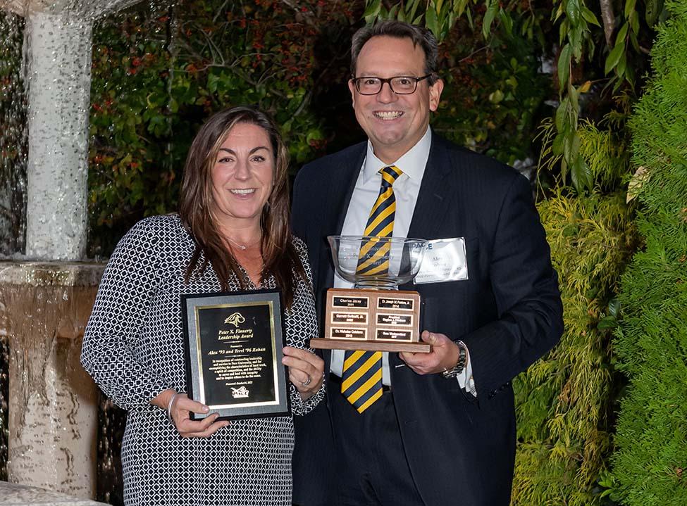 Alex Rohan, wearing a black and white patterned dress, and Terri Rohan, wearing a dark suit and yellow and black tie, hold up the plaques for the Peter X Finnerty Leadership Award 