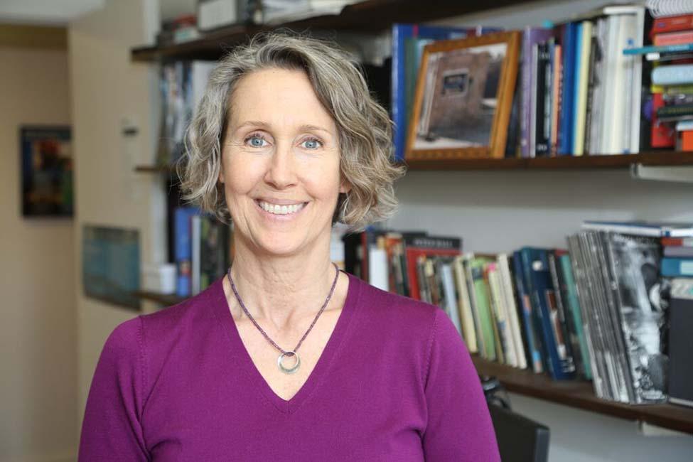 Jane Collins, PhD, wears a purple shirt and stands in front of a full bookshelf
