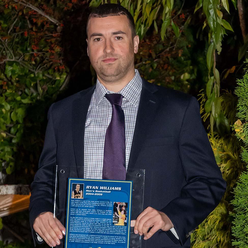 Ryan Williams, wearing a dark suit and purple tie, holds his Hall of Fame plaque