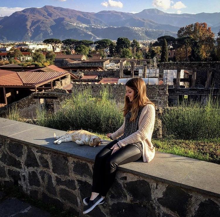 Lubin student Alexis Reed '18 sitting on a stone wall near a village petting a cat during her semester abroad in Italy