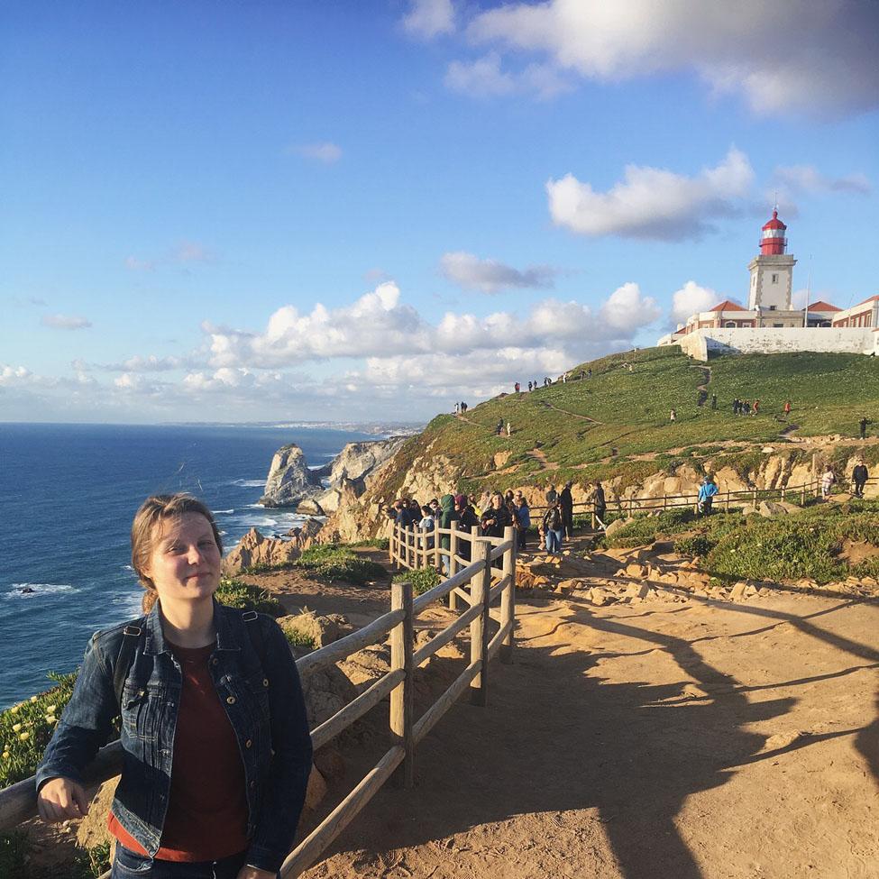 Lubin student Kristina Noreikaite '20 standing on a country road near the sea with a lighthouse on a hill during her semester abroad in Portugal