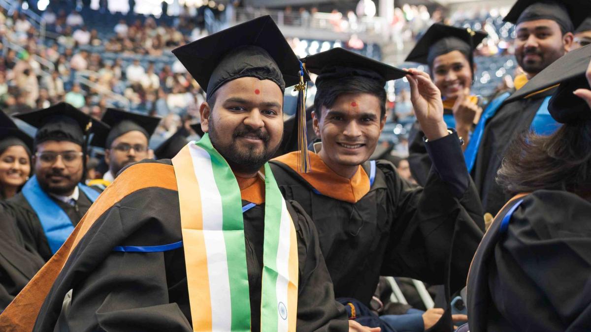 Several students pose in their graduation regalia, one wearing the Indian flag in celebration.