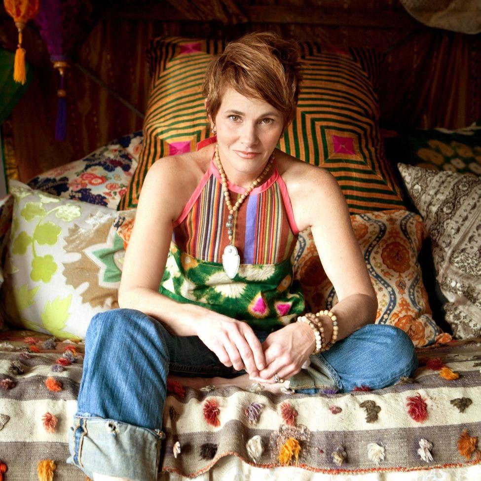A headshot of singer Shawn Colvin sitting on a colorful bed with one leg crossed.