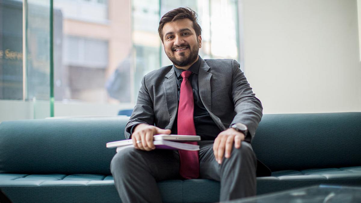 Graduate student sitting on a couch, holding books on his lap and smiling at the camera.