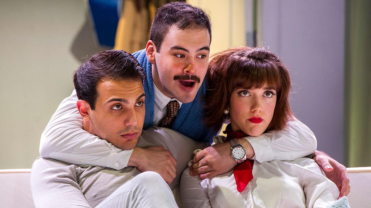 Two actors sitting on a couch with father figure actor behind them with arms around them 
