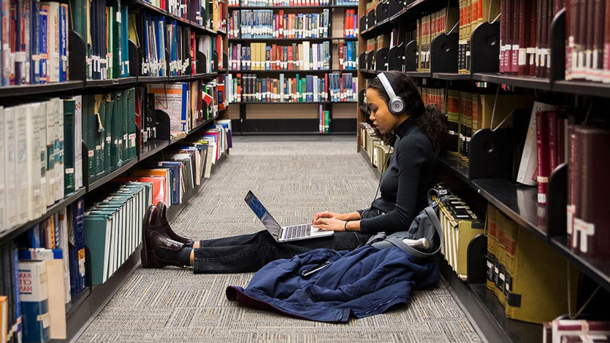 Student sitting with a laptop and headphones between the stacks at the library