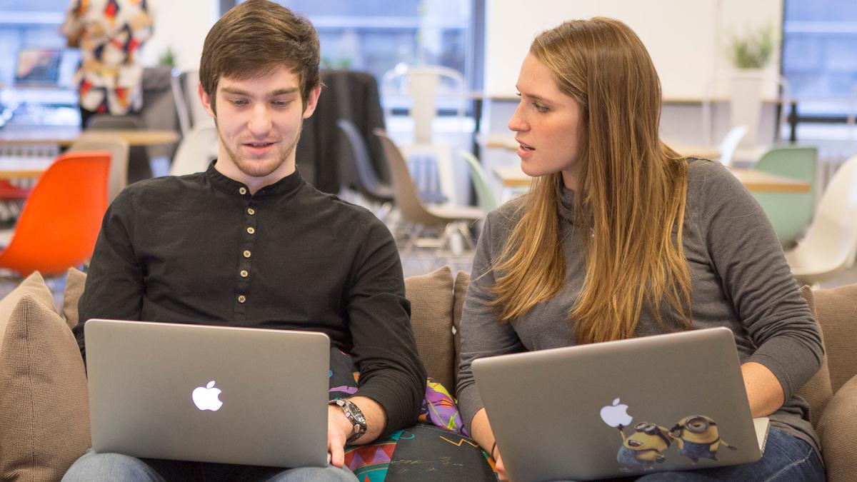 Two students sitting on a couch working on their computers.