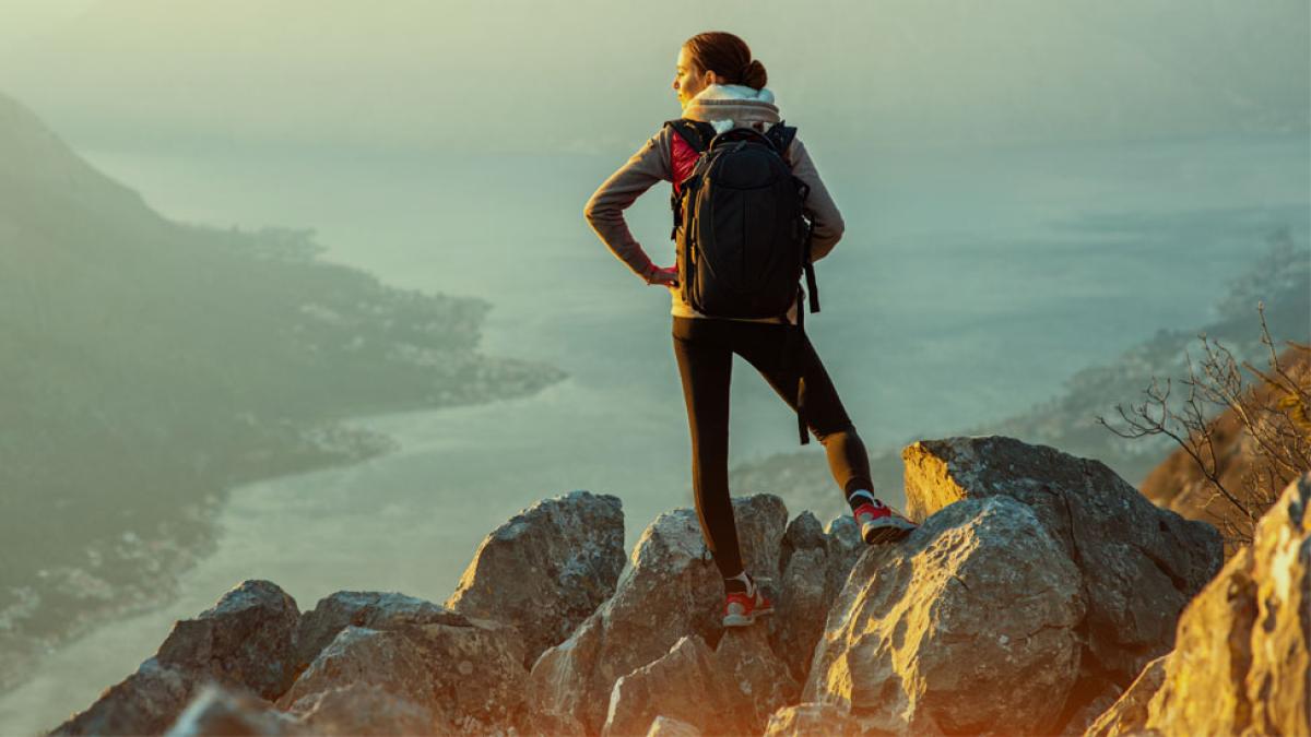 Young traveler with backpack standing on the big stones on the mountain and observing locality.