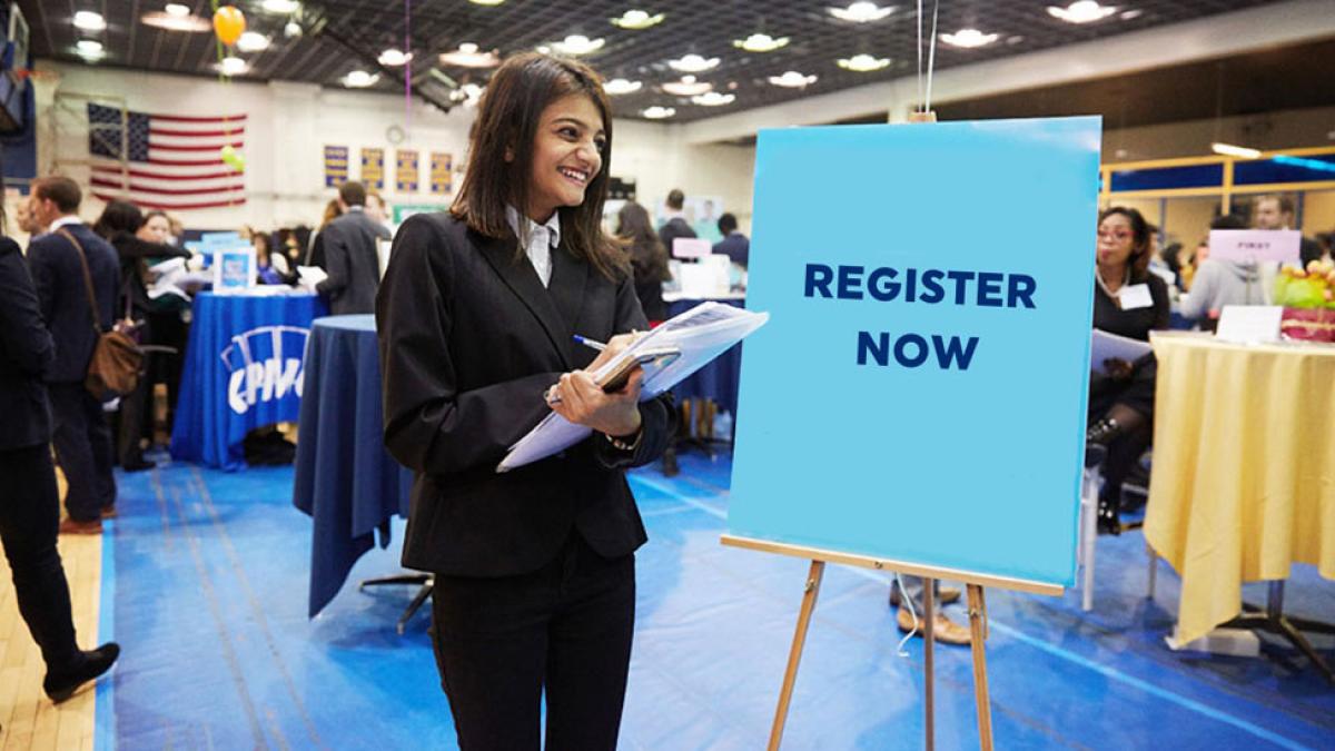 Pace University Career Services employee at a career fair standing in front of a sign that says "Register Here"