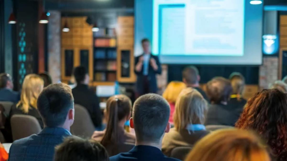 Man in business attire standing at a projection screen in the background presenting to an audience in the foreground