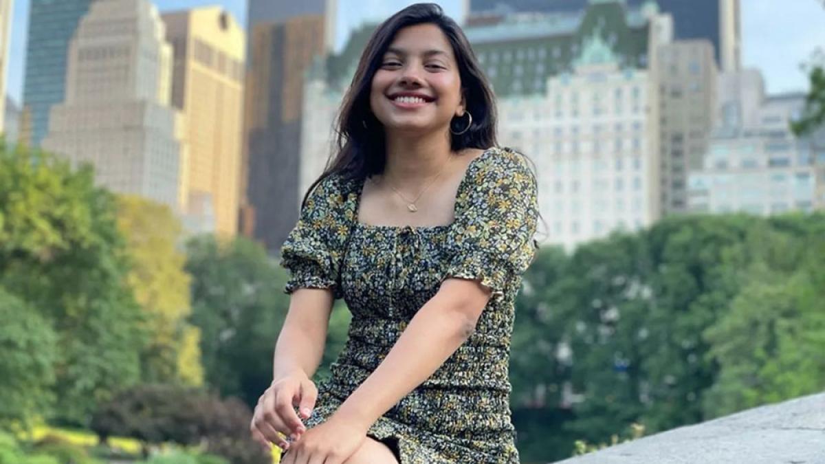 Lubin student Shruti Dhapodkar '23 sitting on a wall in Lower Manhattan in daytime with NYC skyline visible in background