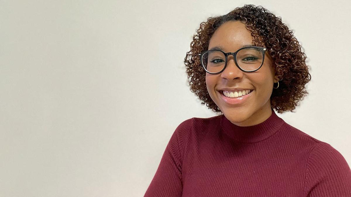 Pace student Mikayla Meachem ’24 wears a red shirt and black glasses and stands in front of a blank background