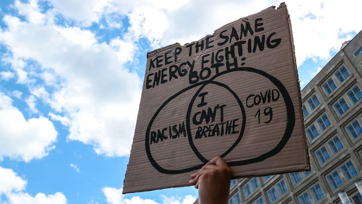 A hand holding up a sign that says "Keep the same engercy fighting for both: Racism  I can't Breathe COVID 19"