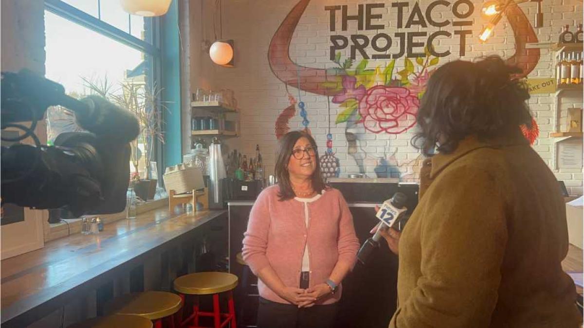 Pace Women's Justice Center ED Cindy Kanusher is seen being interviewed by News12's Carol Wilkinson in front of The Taco Project's mural at their Yonkers location with a camera to the left.