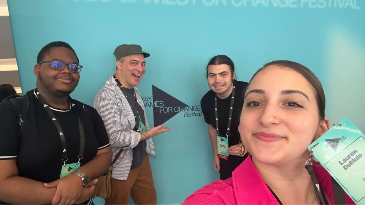 Three Seidenberg students and professor Carmine Guida posing for a photo in front of a banner at the Games for Change Festival.