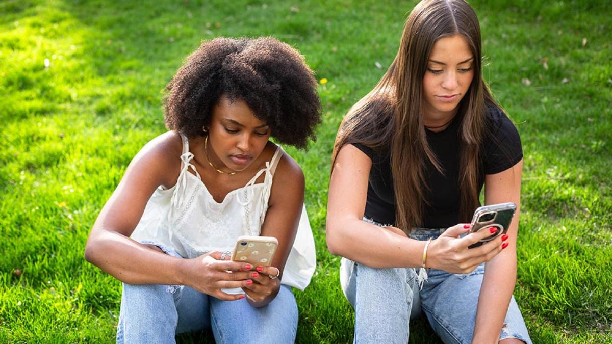 Two young women sitting on the grass and looking at their smartphones.