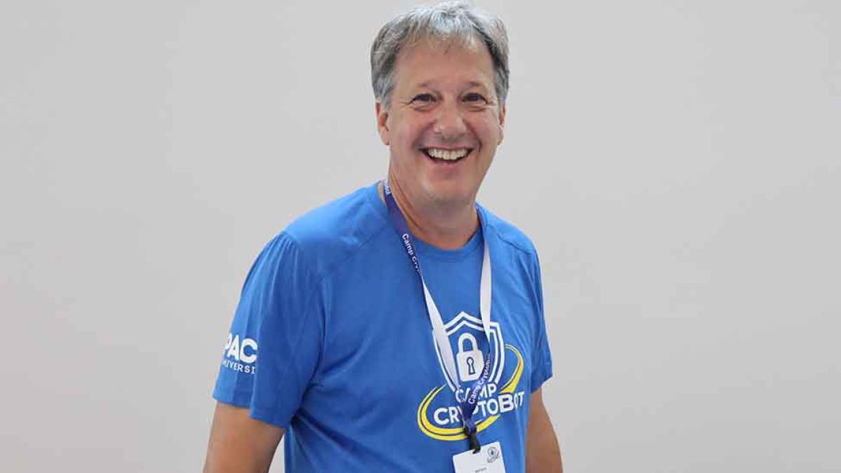 Pace University professor Matt Ganis smiling in a blue shirt against a gray wall