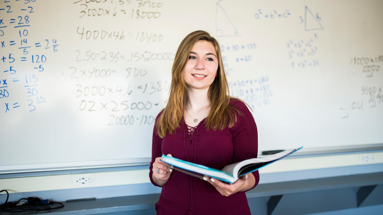 Student standing in front of the board holding a book.