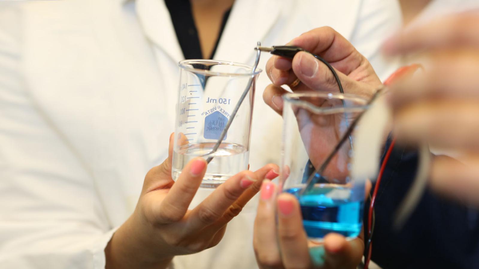 Two students mixing chemicals, one blue and one clear, in wide beakers 