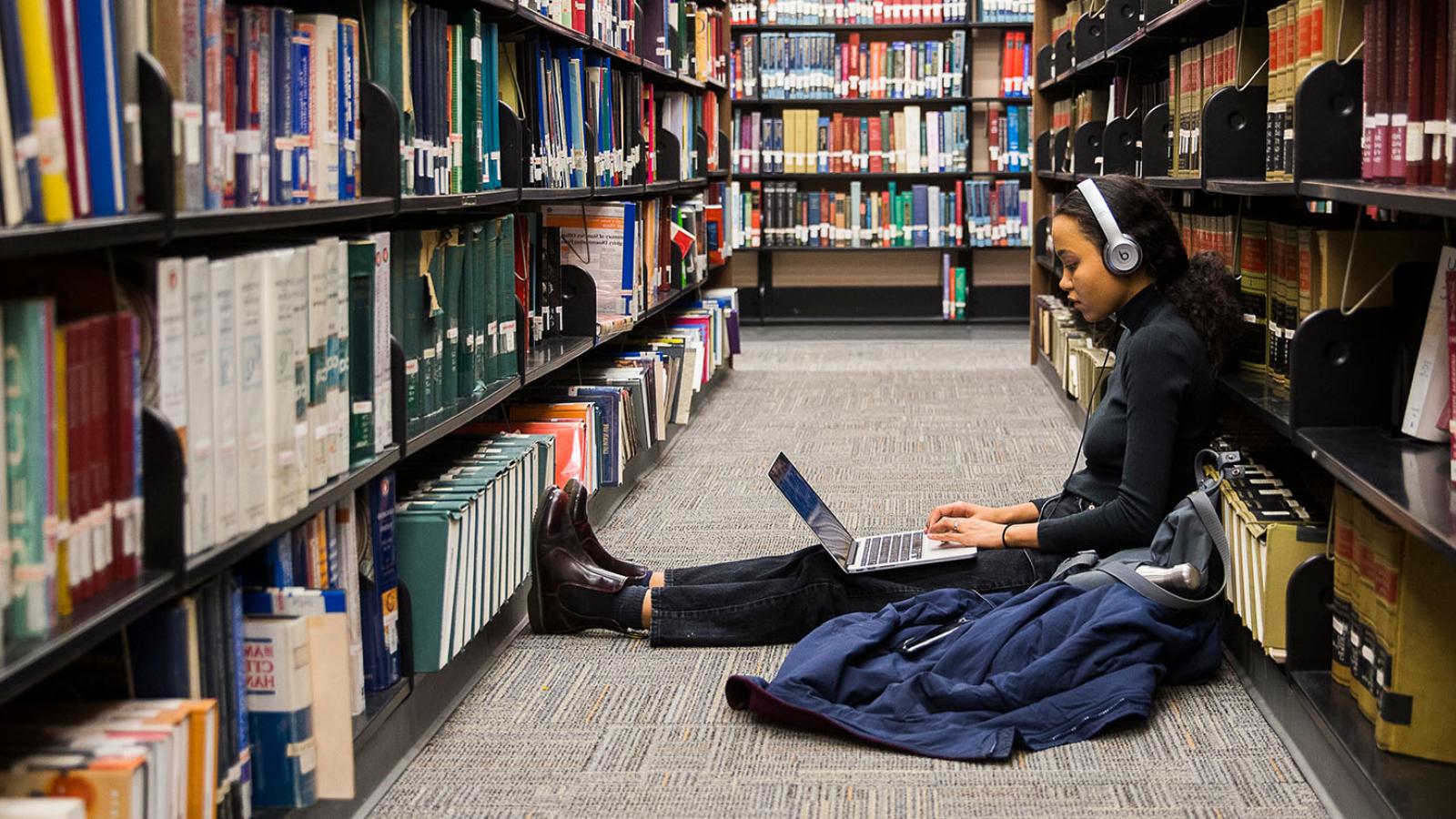 Student sitting between two library bookshelves on laptop wearing headphones