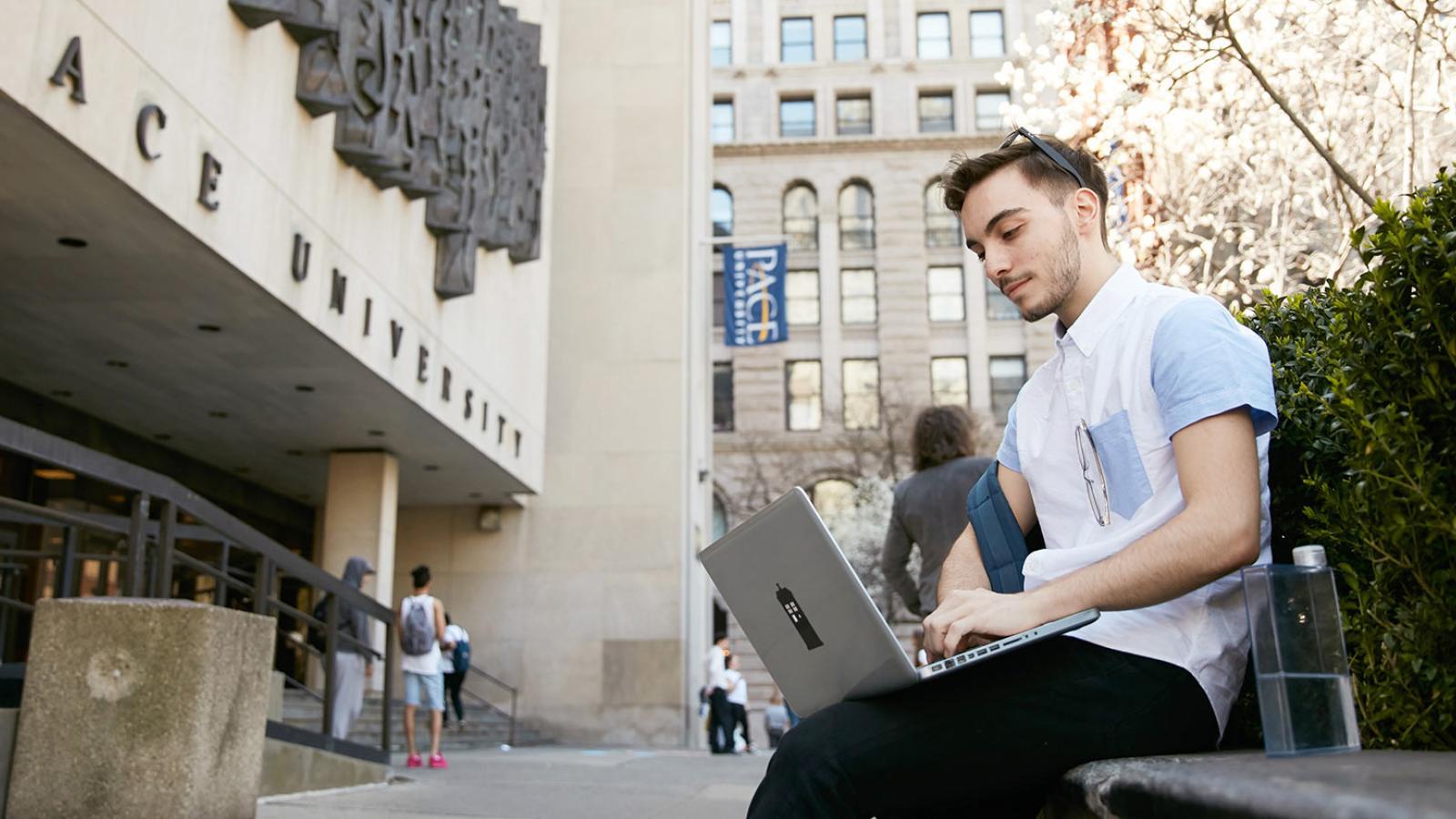 Student sitting in front of 1 Pace Plaza looking at his laptop.