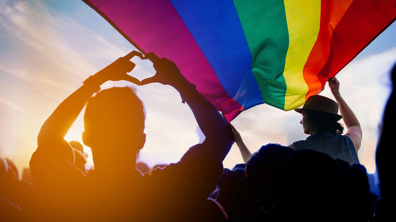 A man makes a heart hand gesture in front of a pride flag