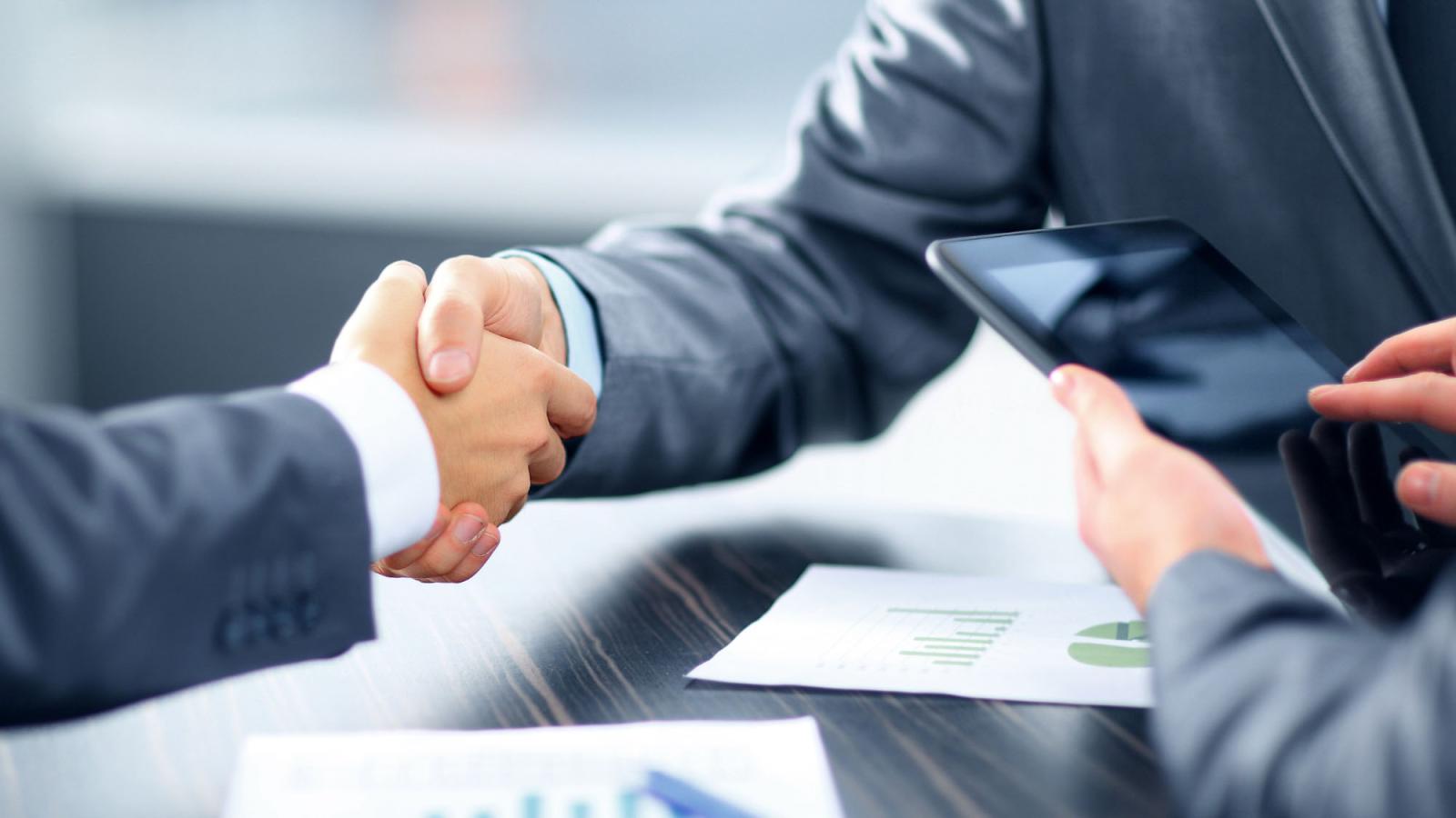 two men in business attire shaking hands near a third person holding a tablet