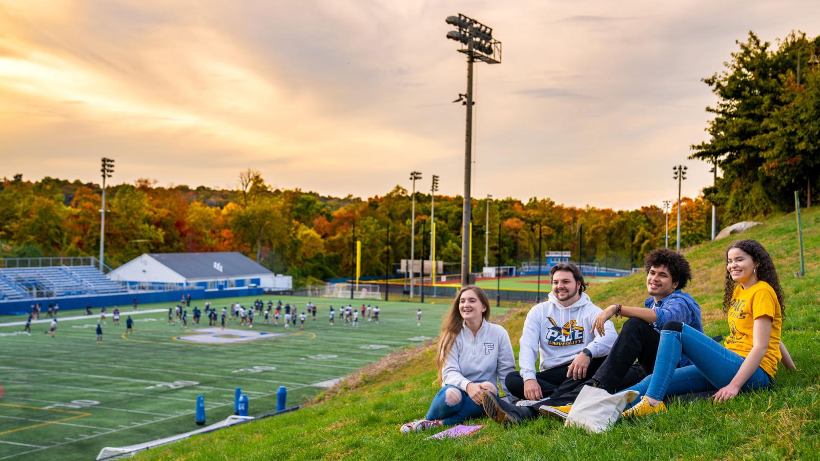 Pace students sit on a grassy hill next to the football field on the Westchester campus