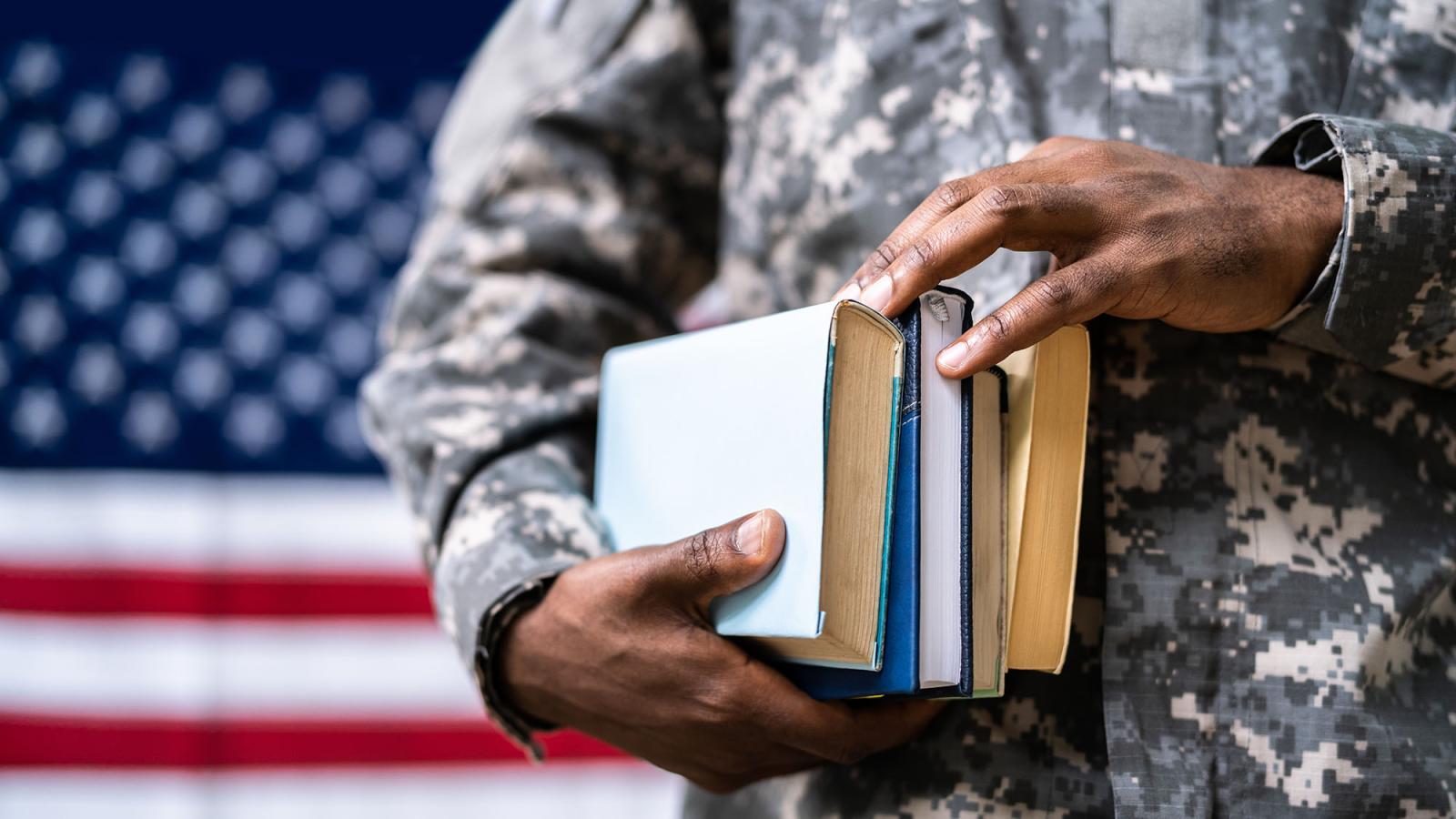 A man wearing a military uniform stands in front of an American flag while holding several books