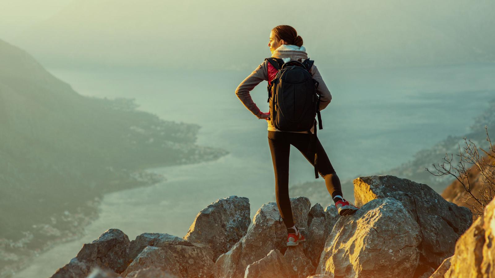 Young traveler with backpack standing on the big stones on the mountain and observing locality.