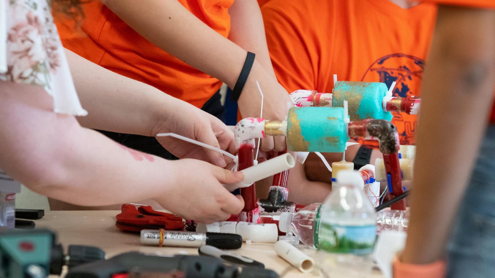 A close up of a robot built using plastic and metal pipes with several pairs of hands working on constructing it.