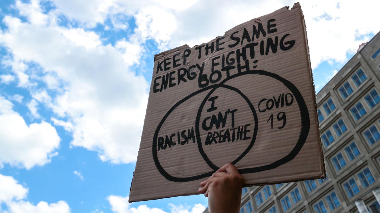 A hand holding up a sign that says "Keep the same engercy fighting for both: Racism I can't Breathe COVID 19"