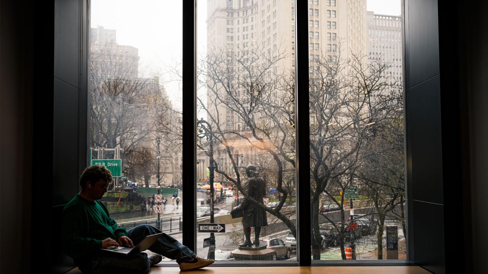 A Pace student in shadow looking out the window of onto City Hall Park.