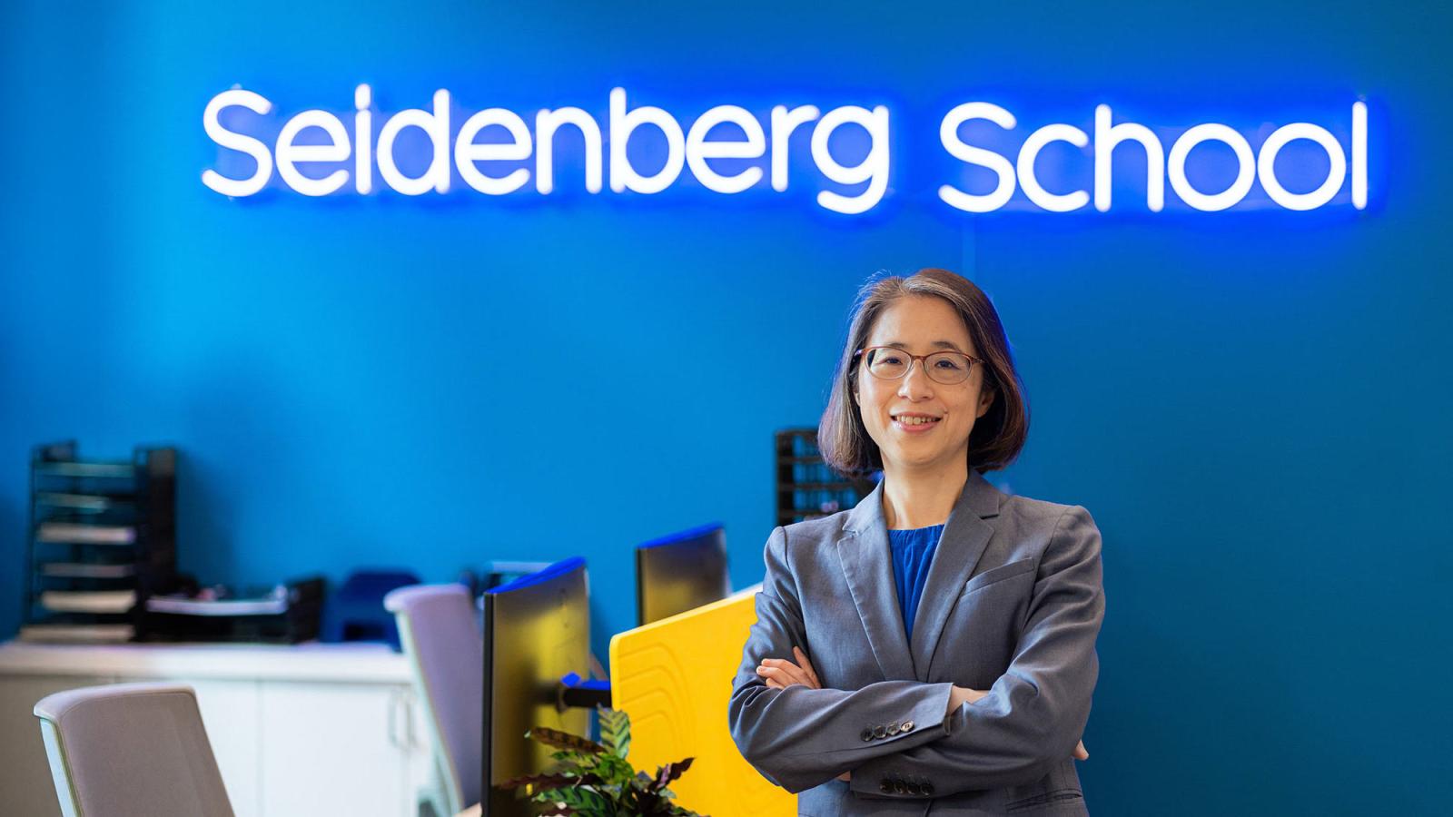 Interim Dean of Seidenberg Li-Chou Chen stands in front of a blue neon sign that says Seidenberg School