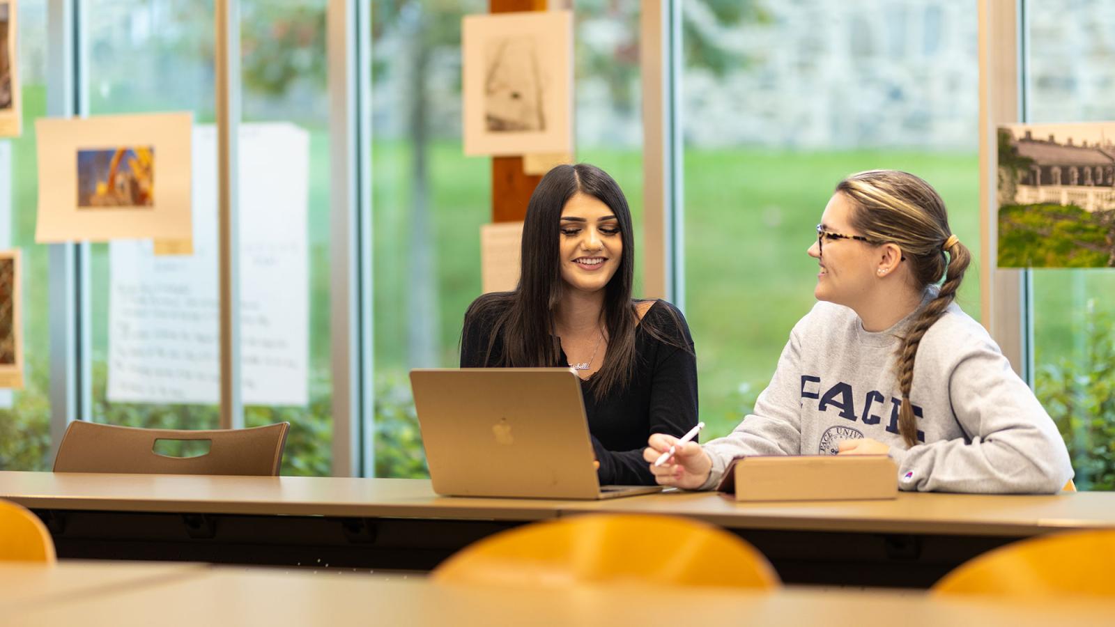 Two Pace University students sitting at a desk, working on a laptop.