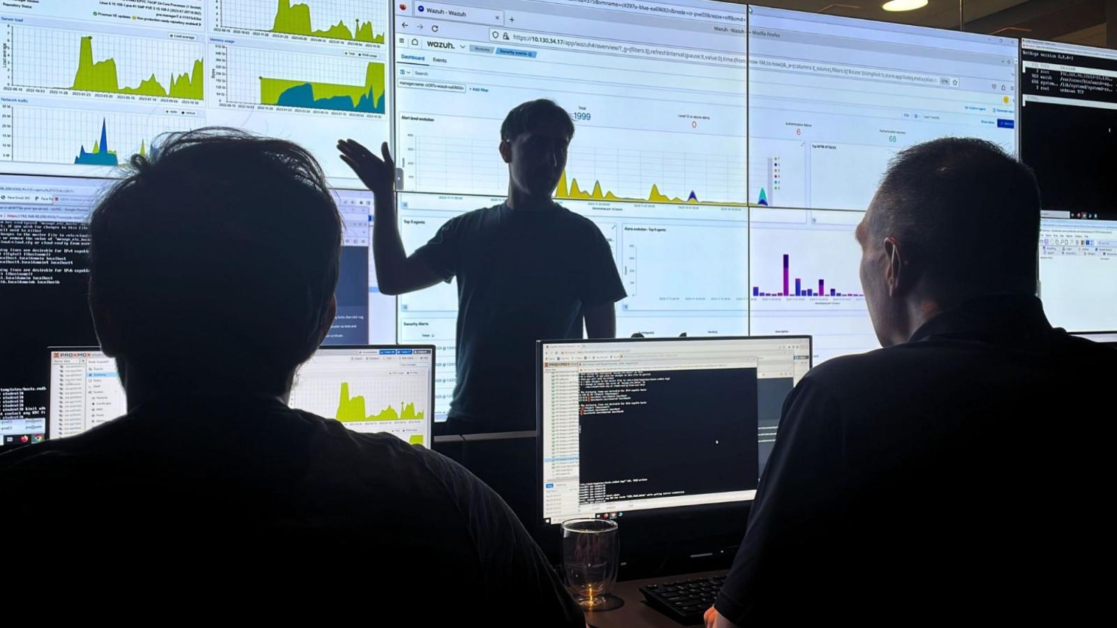 An image of the Seidenberg Cyber Range, in which a student is standing in front of its large screen full of data, explaining the data to another student and faculty member.