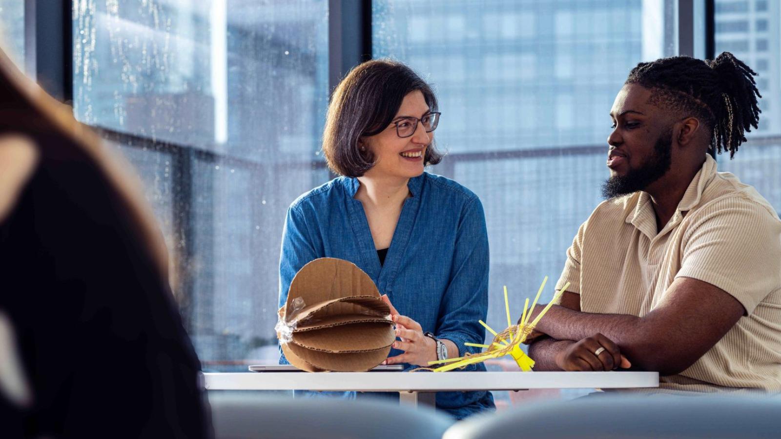 Pace Seidenberg professor and director of the NYC Design Factory Andreea Cotoranu working on a cardboard prototype with a Seidenberg student, with the NYC skyline visible in the window behind them.