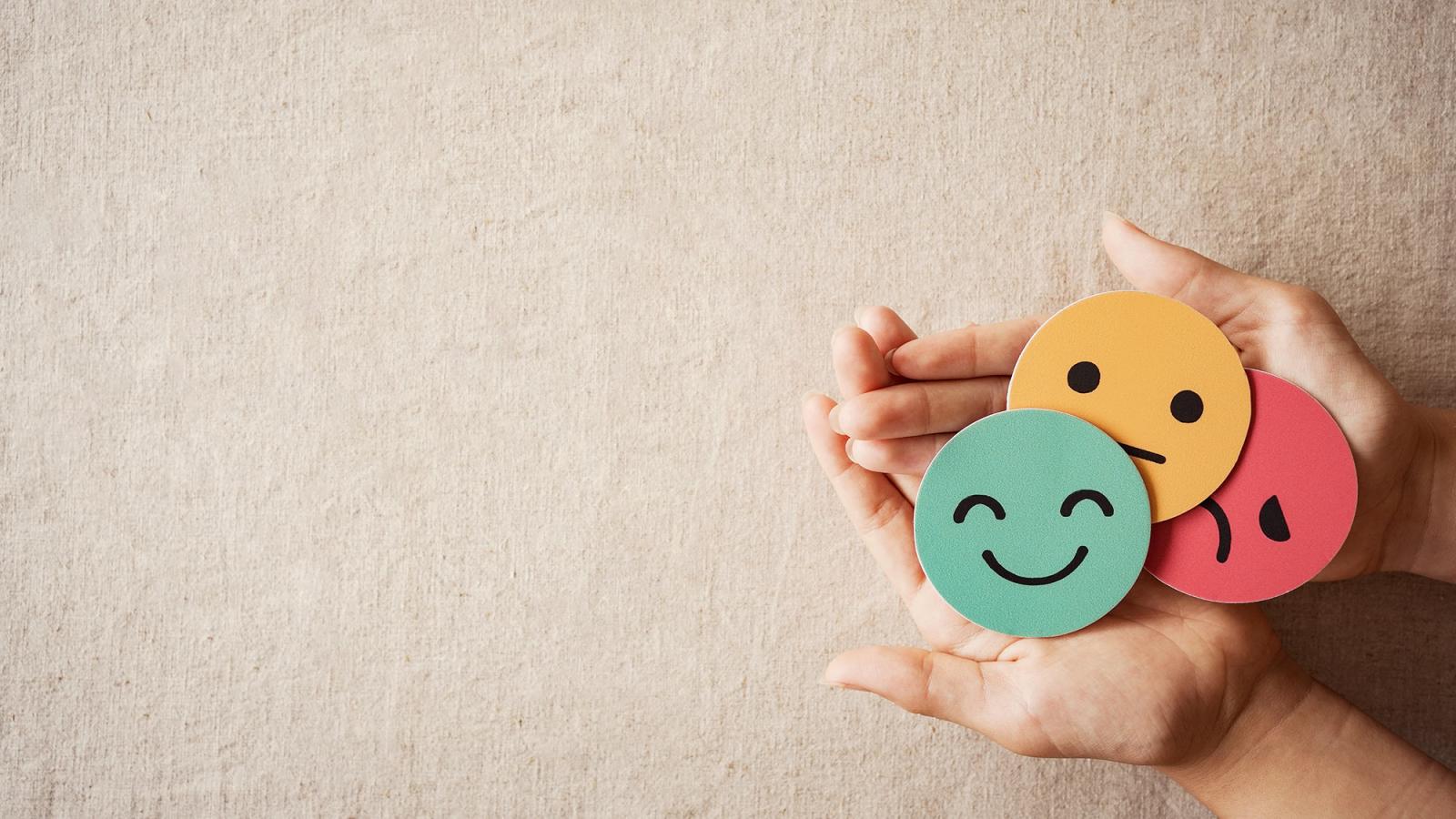 hands set against a beige background hold smiley faces made out of construction paper
