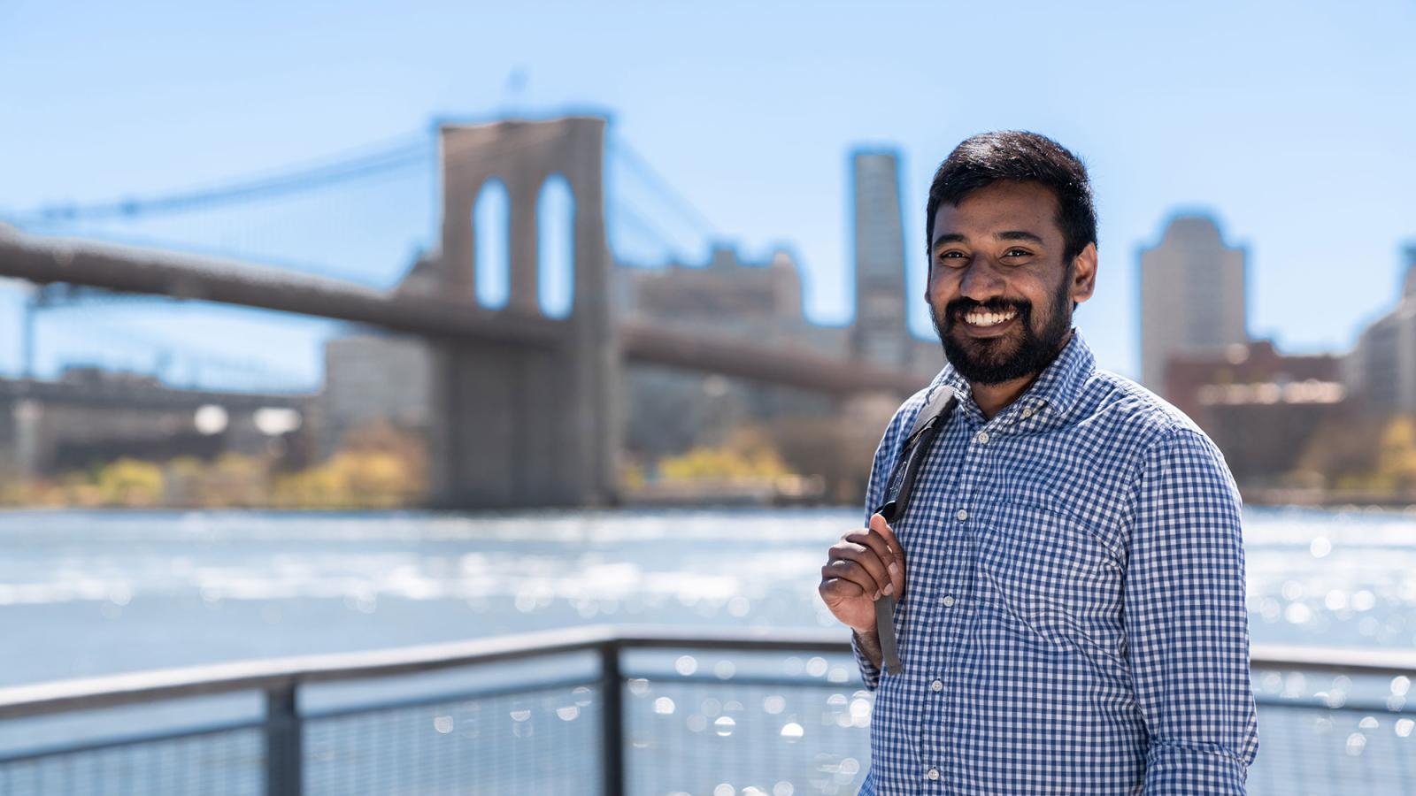 Man, satanding in front of the Brooklyn bridge on a sunny day, smiling at the camera, holding a backpack.