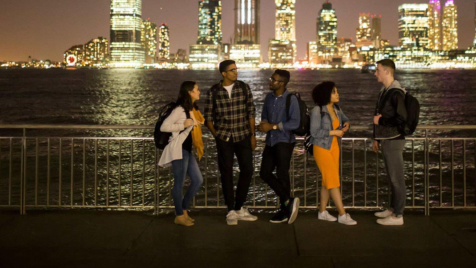 Five students at night looking at the NYC skyline over the water