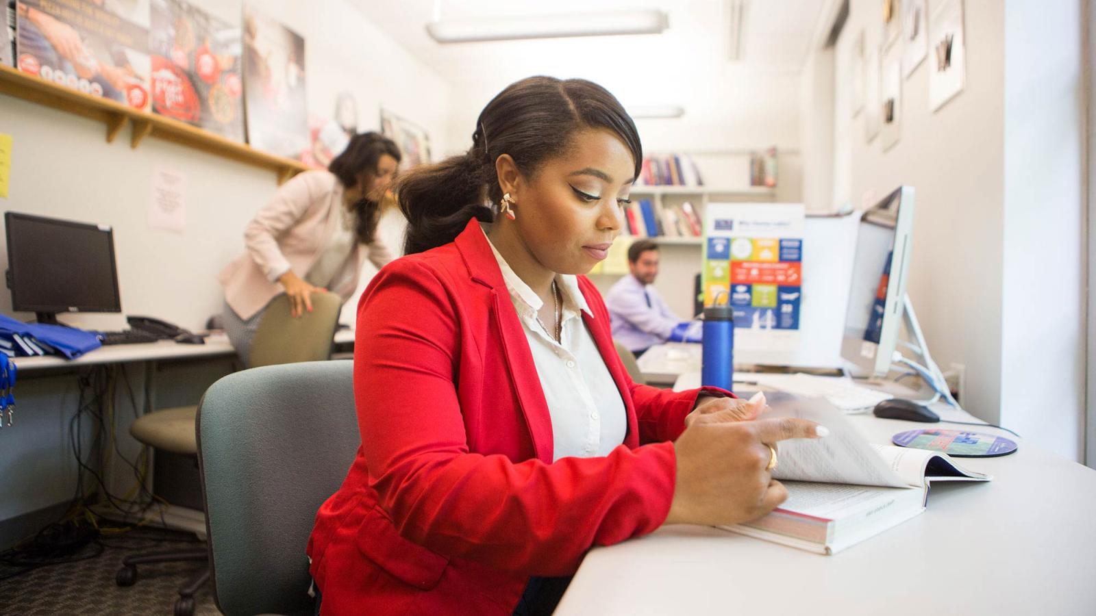 Woman dressed in business attire sitting at a desk in front of a computer, looking at a book.