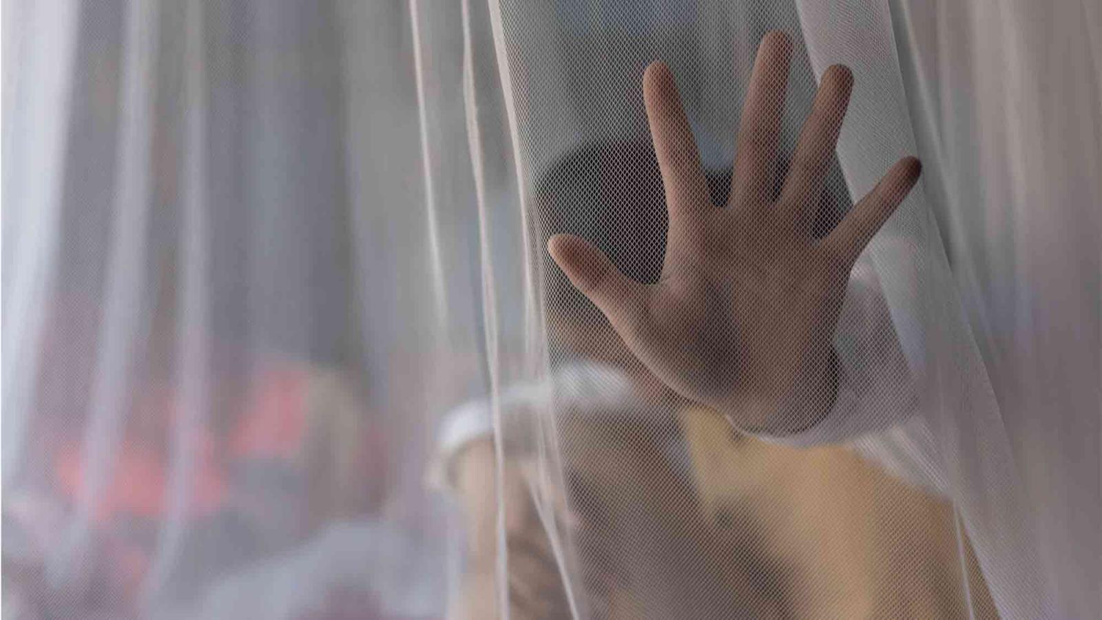 A child’s hand presses against a sheer curtain, partially obscuring their face behind the fabric.