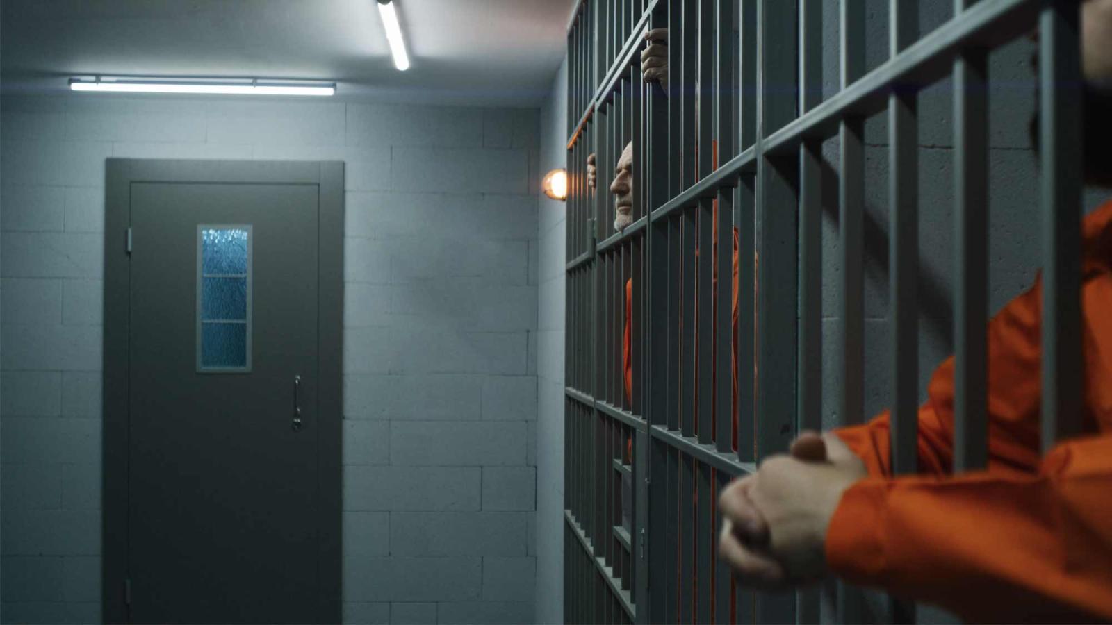 Elderly prisoner in orange uniform stretches fingers, leans on metal bars. Criminal serves imprisonment term for crime in prison cell. Tired inmate stands behind bars in jail or detention center.