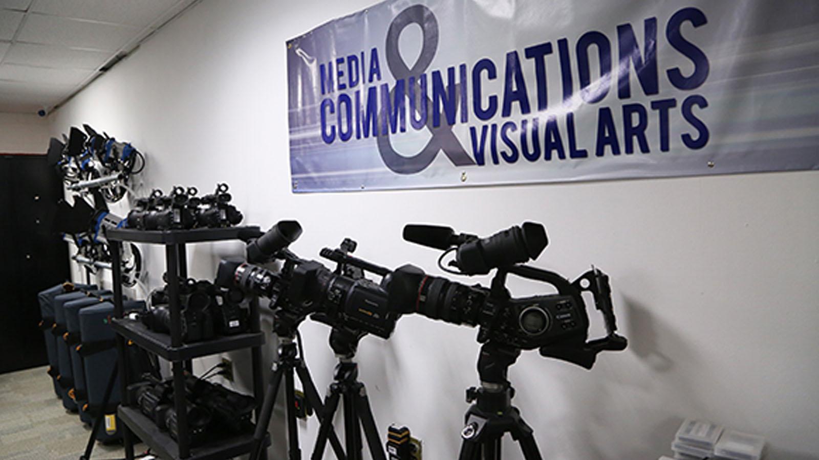 Camera equipment and tripod storage with a Media, Communications, and Visual Arts department banner