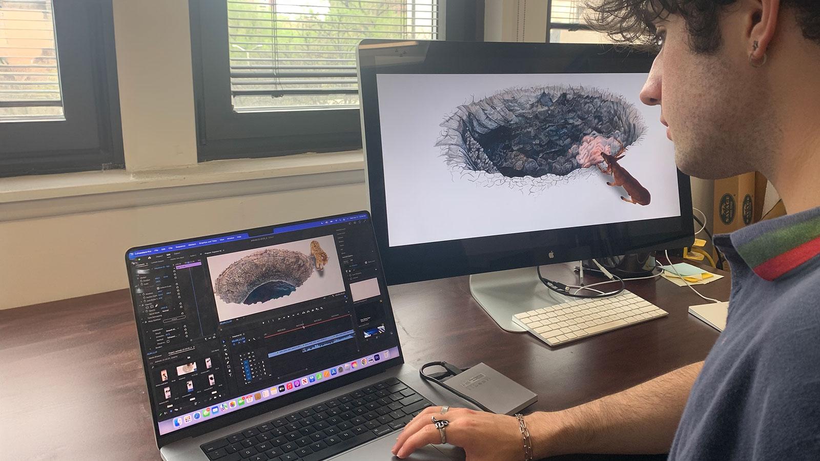 Pace University student Ben Pfiefer working on artwork on a desktop and laptop for Art professor Jillian McDonald's DUMBO Projection Project art exhibition