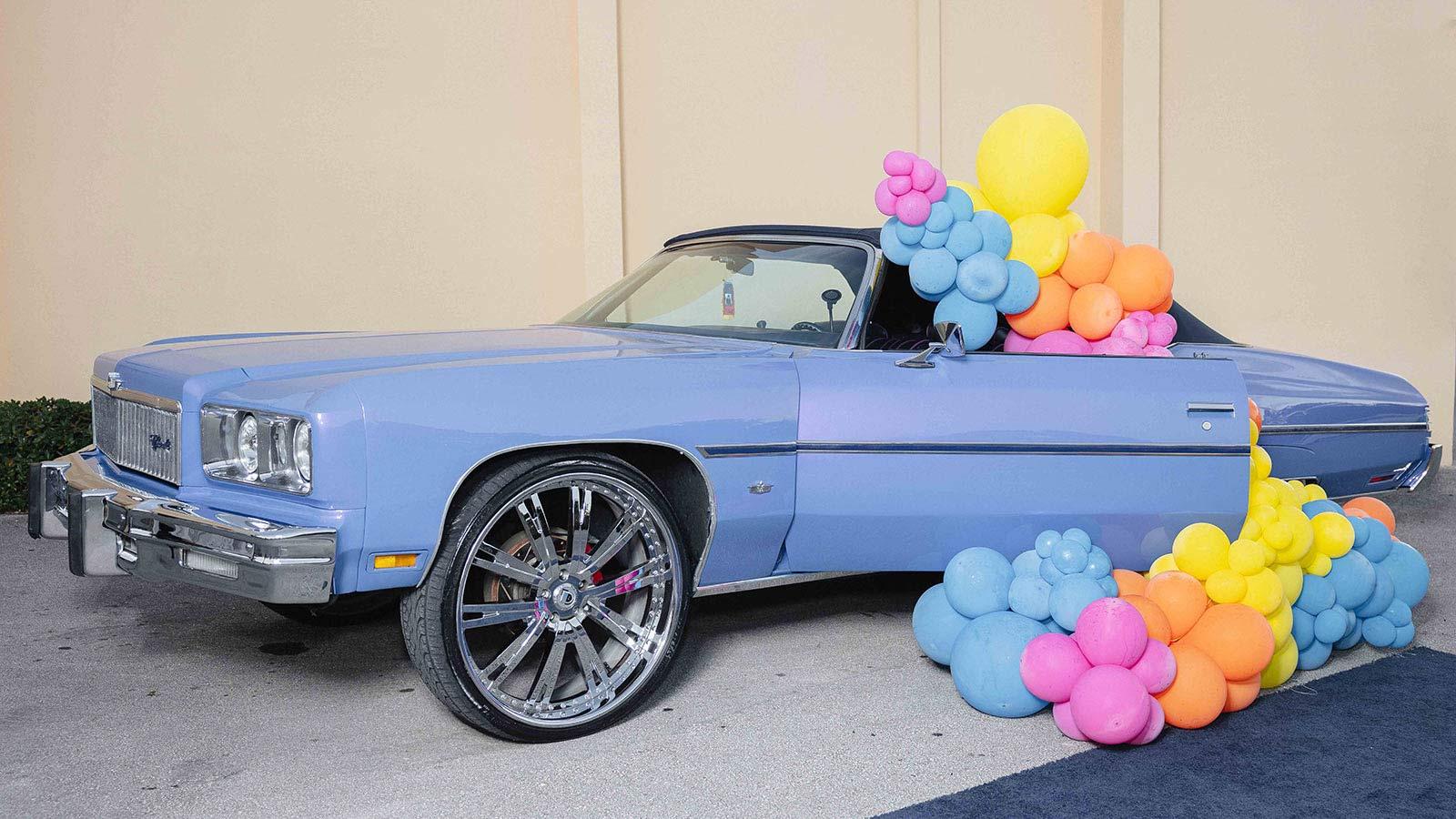 A colorful balloon sculpture designed by a Pace University student surrounds a blue car.