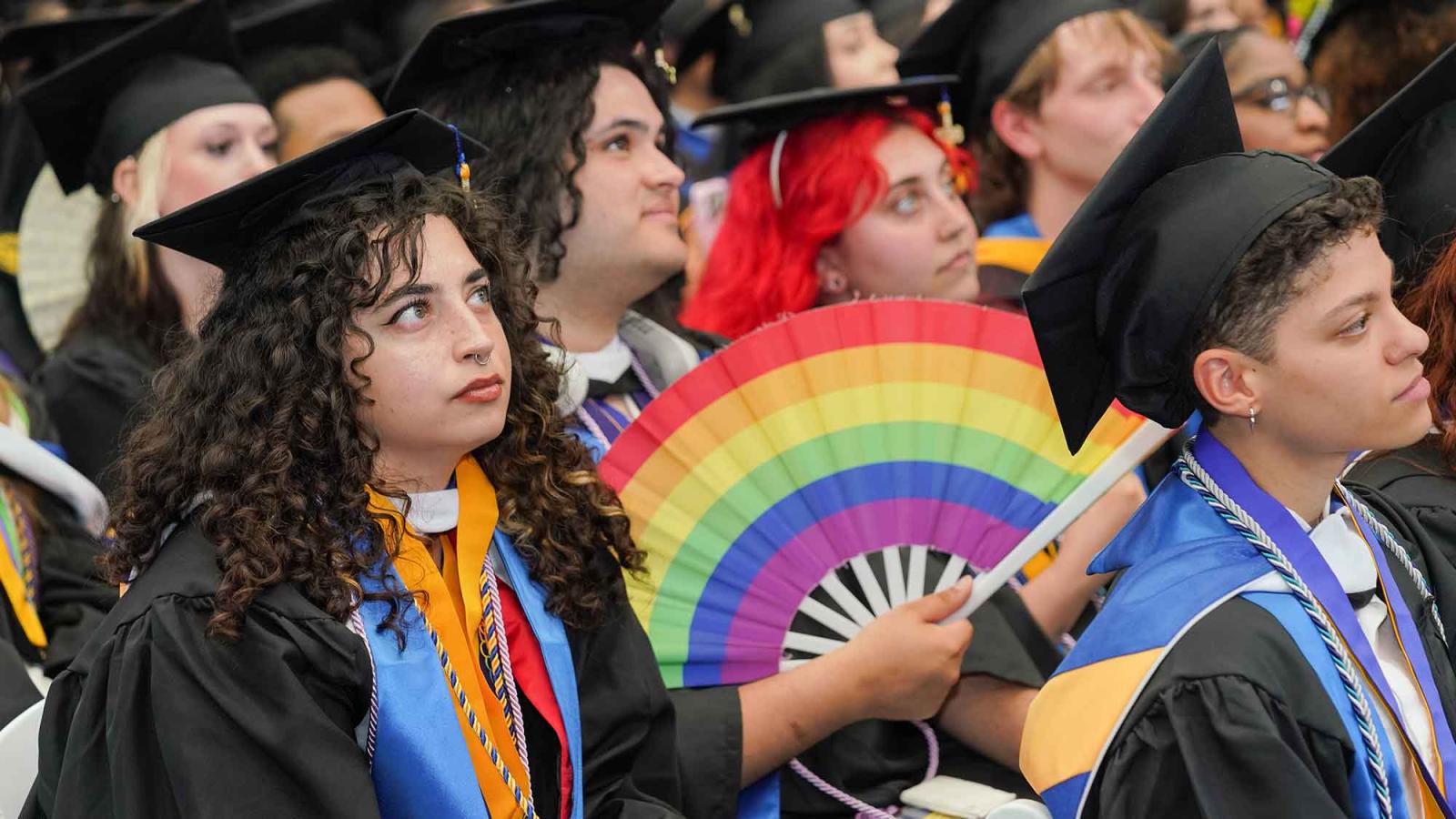 Pace University graduates sit in the crowd, waving a rainbow fan