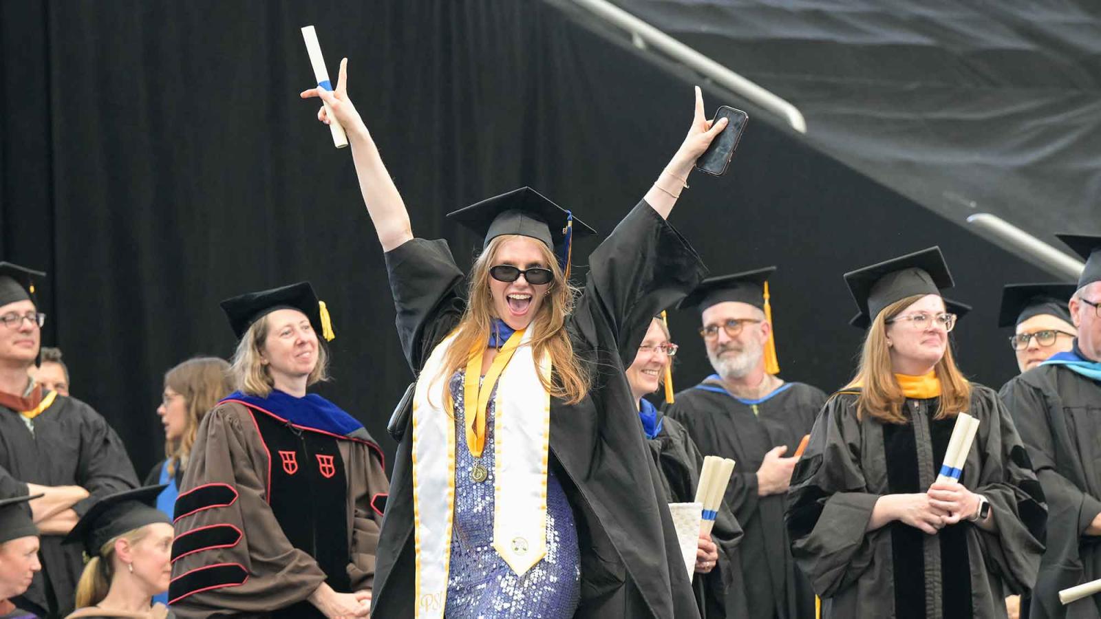 A Pace University graduate wearing sunglasses throws her arms up in the air as she crosses the stage